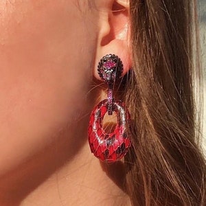 Puede incluir: Pendientes de piel de serpiente con estampado rojo y negro, con un círculo colgante y una pequeña piedra roja.