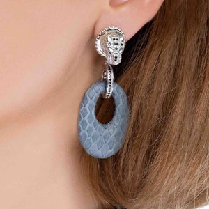 Op de afbeelding: Zilveren oorbellen met een getextureerde grijze cirkel en een zilveren dierenkopbedel met zwarte stenen.