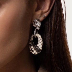 May include: A close-up of a woman's face wearing a pair of silver earrings with a white and black snakeskin pattern. The earrings feature a small crystal detail at the top.