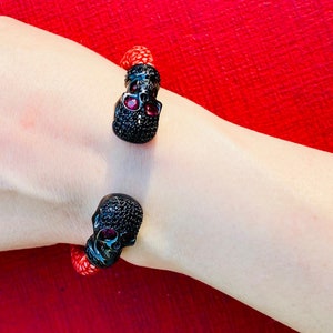 May include: A black skull bracelet with red gemstone eyes. The bracelet is made of red leather and has a skull charm on each end.
