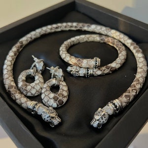 Könnte beinhalten: Ein dreiteiliges Schmuckset mit Schlangenmotiv. Halskette und Armband sind aus weißem Leder mit schwarzen Akzenten. Die Ohrringe sind rund und haben ein weißes Lederdesign mit schwarzen Akzenten.