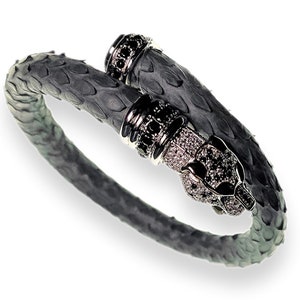 以下が含まれることがあります： シルバーのアクセントが付いたグレーのヘビ革カフブレスレット。ブレスレットは、黒と透明の宝石が付いたパンサーヘッドのデザインが特徴です。スタイリッシュなアクセサリーです。