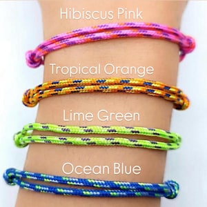 May include: Four colorful rope bracelets are displayed, each with a different color and name: Hibiscus Pink, Tropical Orange, Lime Green, and Ocean Blue. The bracelets are knotted at the ends and are worn on a wrist.