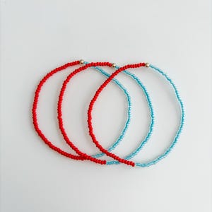 May include: Three circular beaded bracelets arranged in an overlapping pattern. Two bracelets are composed of small, vibrant red beads, while the third is made of light blue beads. Each bracelet features a single, small, metallic gold-tone bead.