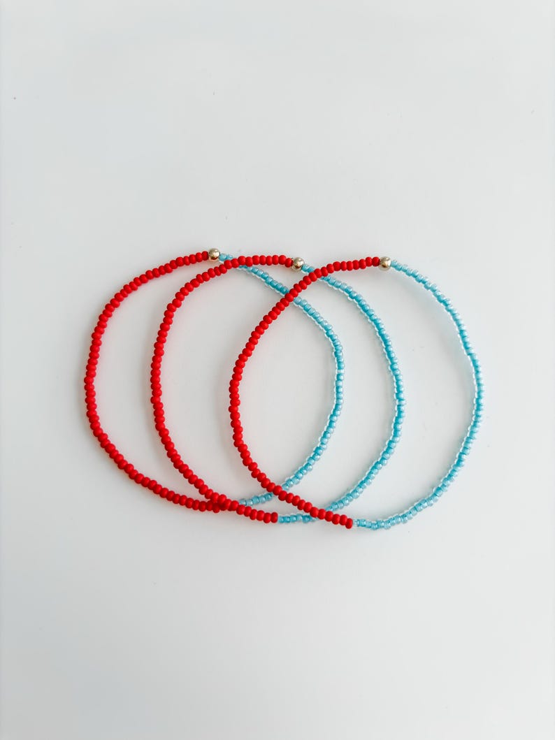 May include: Three circular beaded bracelets arranged in an overlapping pattern. Two bracelets are composed of small, vibrant red beads, while the third is made of light blue beads. Each bracelet features a single, small, metallic gold-tone bead.