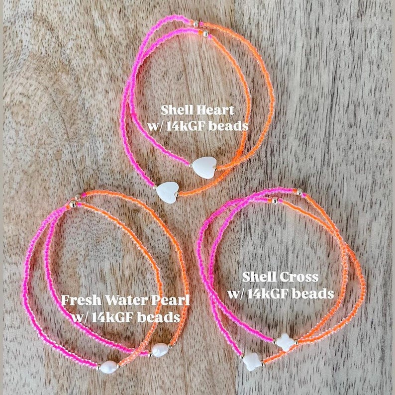 May include: Three bracelets featuring neon pink and orange beaded strands with 14k gold-filled beads. One bracelet has white shell hearts, one has white freshwater pearls, and one has white shell crosses, labeled Shell Heart, Fresh Water Pearl, and Shell Cross.