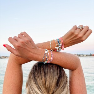 May include: A collection of layered beaded bracelets worn on both wrists. The bracelets feature small colorful glass beads in shades of pink, blue, orange, and green, accented with small white heart-shaped charms and metallic silver spacers.