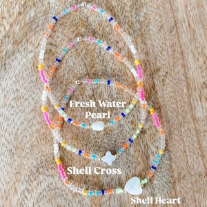 May include: Three beaded bracelets featuring colorful glass beads in shades of pink, orange, yellow, blue, and white. The bracelets include charms labeled Fresh Water Pearl, Shell Cross, and Shell Heart, each featuring a white shell or pearl.