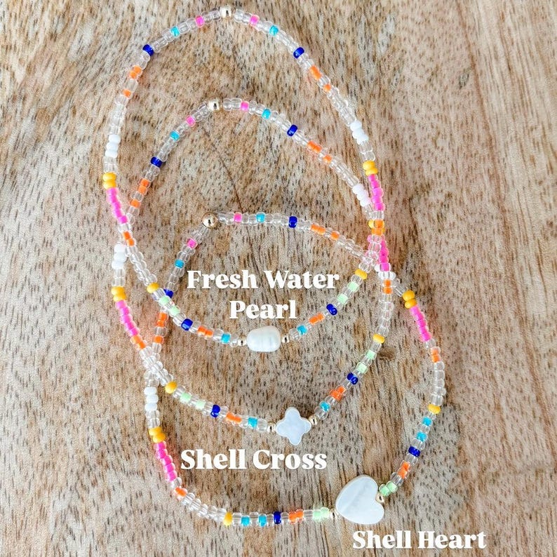 May include: Three beaded bracelets featuring colorful glass beads in shades of pink, orange, yellow, blue, and white. The bracelets include charms labeled Fresh Water Pearl, Shell Cross, and Shell Heart, each featuring a white shell or pearl.