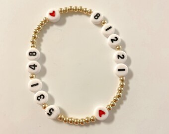 May include: A gold beaded bracelet with white beads that have black numbers on them. The numbers on the beads spell out "12181348" and there are two red heart beads.