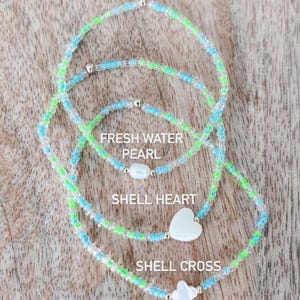 May include: Three beaded bracelets made of small light blue, lime green, and clear glass beads. The bracelets feature a central white freshwater pearl, a white shell heart, and a white shell cross, respectively, with the text labels included.