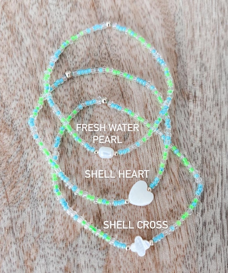 May include: Three beaded bracelets made of small light blue, lime green, and clear glass beads. The bracelets feature a central white freshwater pearl, a white shell heart, and a white shell cross, respectively, with the text labels included.