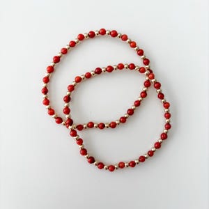May include: Two circular beaded bracelets featuring an alternating pattern of small, round, vibrant red beads and polished metallic gold-tone beads. The bracelets are arranged in an overlapping circular formation against a plain white background.