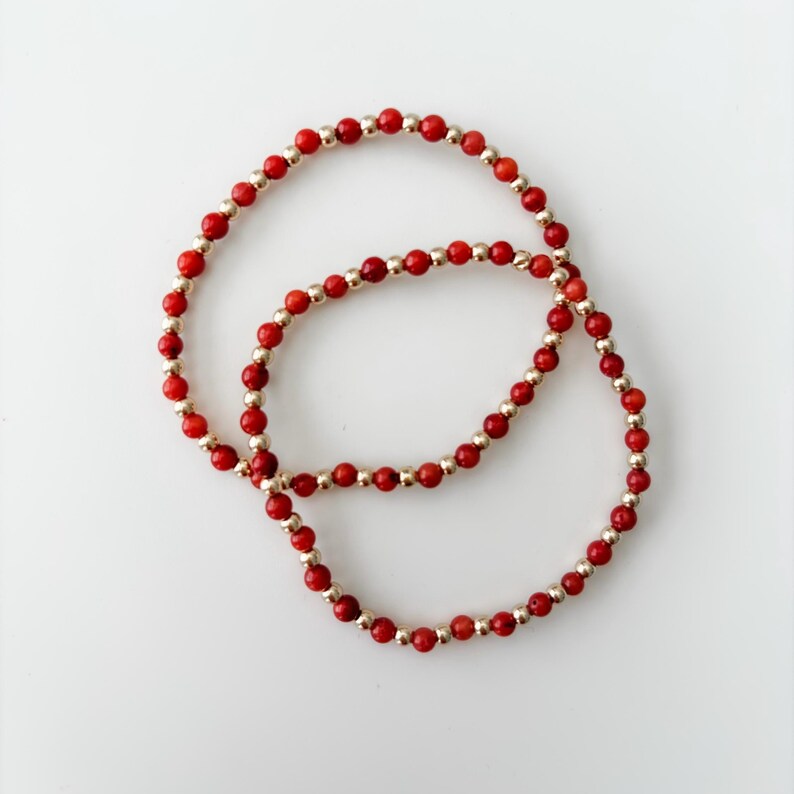 May include: Two circular beaded bracelets featuring an alternating pattern of small, round, vibrant red beads and polished metallic gold-tone beads. The bracelets are arranged in an overlapping circular formation against a plain white background.