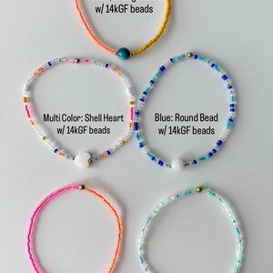 May include: Five beaded bracelets featuring 14k gold-filled beads. The bracelets include a yellow and orange strand with a blue bead, a multi-color strand with a shell heart, a blue strand with a round bead, a pink and orange strand with a pearl, and a green and blue strand with a shell cross.
