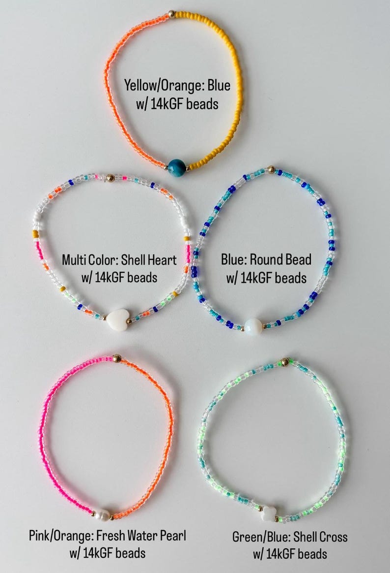 May include: Five beaded bracelets featuring 14k gold-filled beads. The bracelets include a yellow and orange strand with a blue bead, a multi-color strand with a shell heart, a blue strand with a round bead, a pink and orange strand with a pearl, and a green and blue strand with a shell cross.