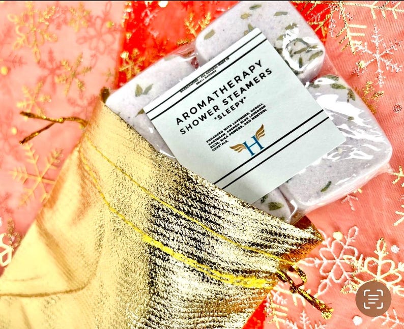K&ouml;nnte beinhalten: Ein Set mit vier Aromatherapie-Duschdampfern in einer goldenen Tasche. Die Duschdampfer sind wei&szlig; und haben einen Lavendel-Duft. Das Etikett lautet "Aromatherapy Shower Steamers *Sleepy*"