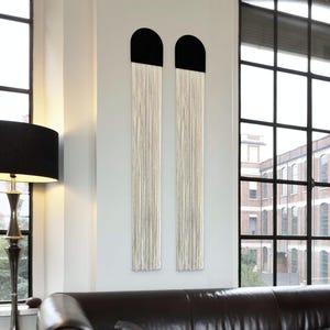 May include: Two vertical wall hangings with a black, arched top and long, cream-colored fringe. The art pieces are mounted on a light-colored wall, with a black lamp and a leather sofa in the foreground. Large windows provide natural light.