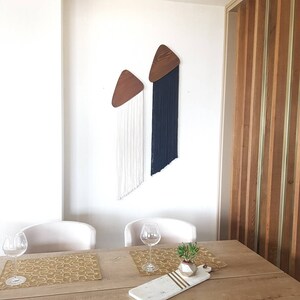 K&ouml;nnte beinhalten: Zwei Wandbeh&auml;nge aus Holz in Dreiecksform mit langen wei&szlig;en und schwarzen Garnstr&auml;ngen, die von unten h&auml;ngen. Die Wandbeh&auml;nge befinden sich an einer wei&szlig;en Wand in einem Esszimmer mit einem Holztisch, zwei St&uuml;hlen und einer kleinen Topfpflanze.