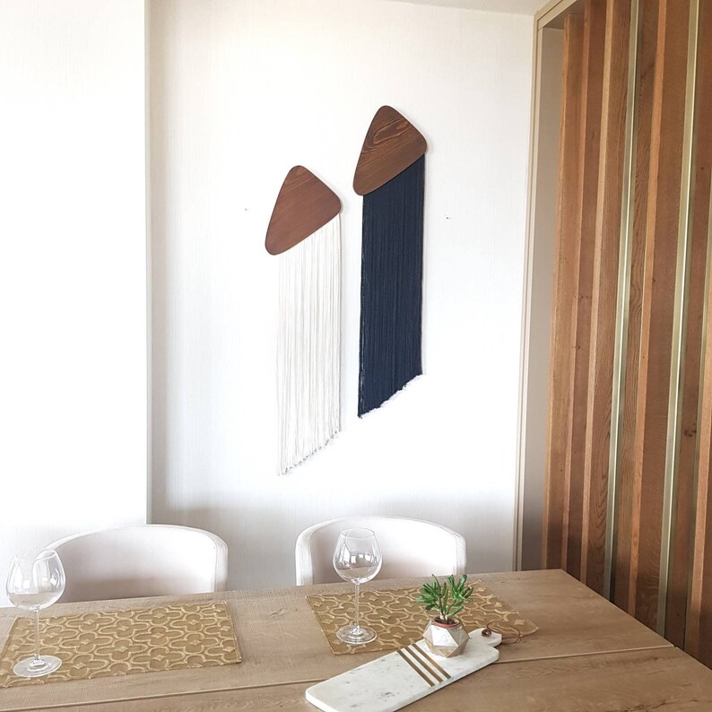 K&ouml;nnte beinhalten: Zwei Wandbeh&auml;nge aus Holz in Dreiecksform mit langen wei&szlig;en und schwarzen Garnstr&auml;ngen, die von unten h&auml;ngen. Die Wandbeh&auml;nge befinden sich an einer wei&szlig;en Wand in einem Esszimmer mit einem Holztisch, zwei St&uuml;hlen und einer kleinen Topfpflanze.