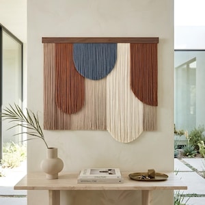 May include: A large, rectangular wall hanging with a wooden frame. The artwork features a design of vertical strands in shades of brown, blue, and cream. The piece is mounted on a neutral-colored wall, creating a modern, minimalist aesthetic.
