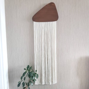 K&ouml;nnte beinhalten: Ein wei&szlig;er Makramee-Wandbehang mit einer dreieckigen Holzform oben.