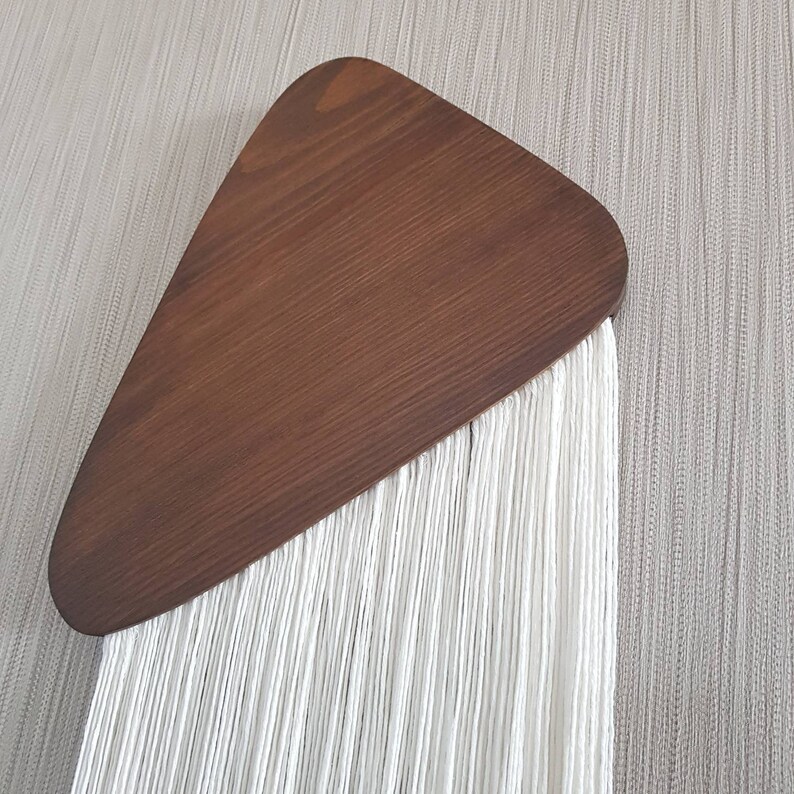 K&ouml;nnte beinhalten: Ein wei&szlig;er Makramee-Wandbehang mit einem braunen Holzdreieck oben.