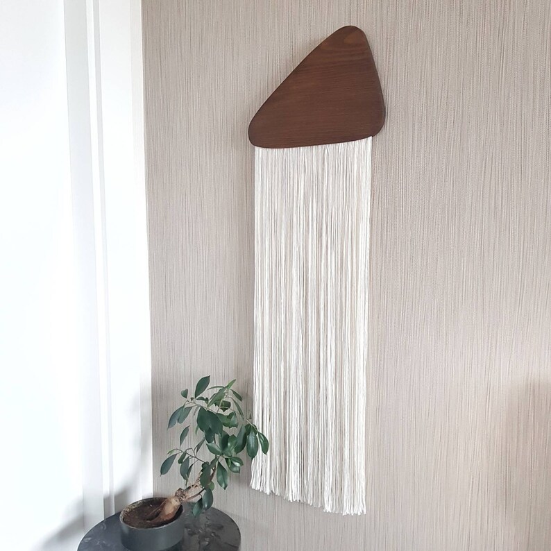 K&ouml;nnte beinhalten: Ein wei&szlig;er Makramee-Wandbehang mit einer dreieckigen Holzform oben. Der Makramee besteht aus d&uuml;nnen wei&szlig;en Schn&uuml;ren und h&auml;ngt an dem Holzdreieck.