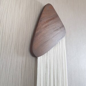 K&ouml;nnte beinhalten: Ein Wandbehang aus wei&szlig;em Garn mit einem braunen Holzdreieck oben.