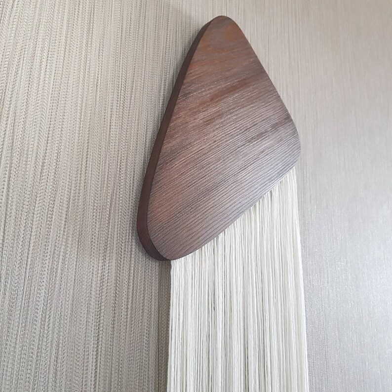 K&ouml;nnte beinhalten: Ein Wandbehang aus wei&szlig;em Garn mit einem braunen Holzdreieck oben.