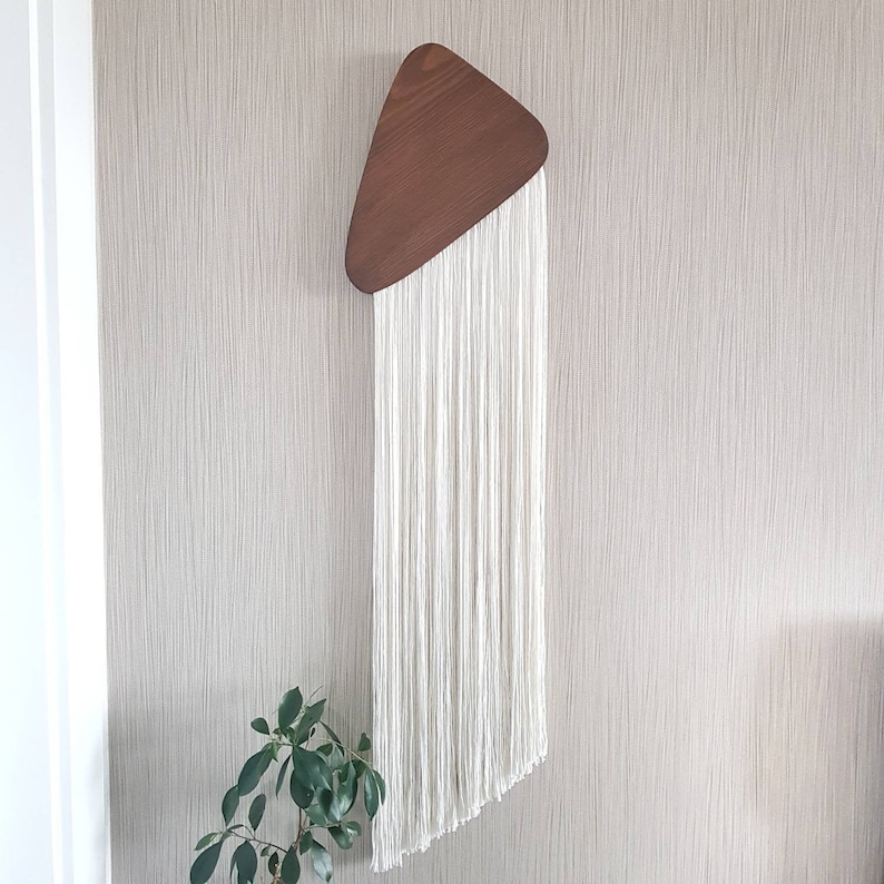 K&ouml;nnte beinhalten: Ein wei&szlig;er Makramee-Wandbehang mit einer dreieckigen Holzform oben.
