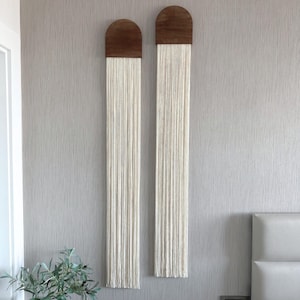 May include: Two wall hangings with a wooden arched top and long, cream-coloured fringe. The wooden tops are a medium brown colour. The hangings are displayed on a textured, light grey wall, adding a touch of bohemian decor.