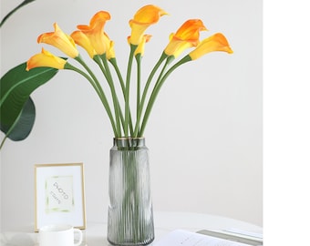 May include: A bouquet of yellow calla lilies in a clear, textured glass vase with a gold rim. The vase is on a white table with a book and a white cup and saucer.