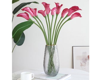 May include: A bouquet of pink calla lilies arranged in a clear glass vase with a gold rim. The vase is sitting on a white table with a book and a white cup and saucer.