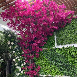 Pode incluir: Uma exibição floral artificial com uma árvore de vibrantes flores magenta, contra paredes de folhagem verde e arbustos de rosas brancas. A cena é iluminada por iluminação superior, criando um ambiente luminoso.
