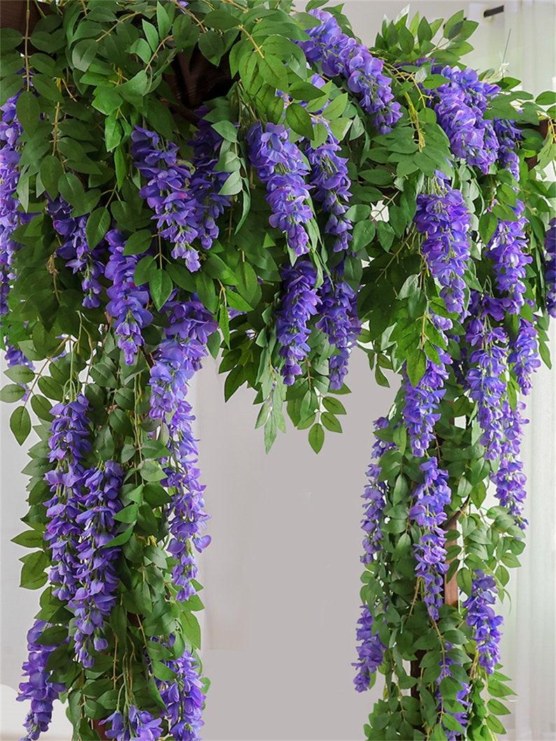 May include: Artificial wisteria garland with cascading purple flowers and green leaves. The wisteria is draped over a wooden structure, creating a lush, floral display. Ideal for weddings, home decor, or event decorations.