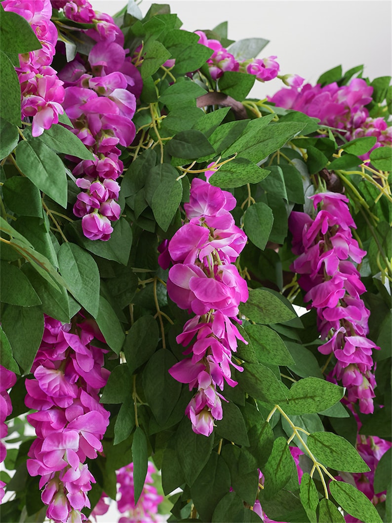 May include: Close-up of artificial wisteria vines with vibrant magenta flowers and lush green leaves. The cascading flowers are in full bloom, creating a dense and colorful display. The image showcases the intricate details of the floral arrangement.