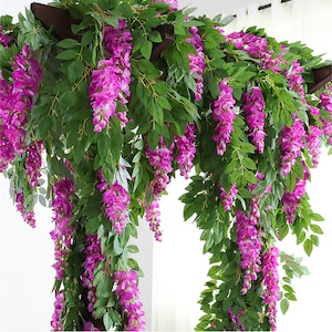 May include: Artificial wisteria garland with cascading purple flowers and green leaves. The garland is draped over a dark brown wooden structure, creating a vibrant and decorative display. Ideal for adding a touch of nature to any space.