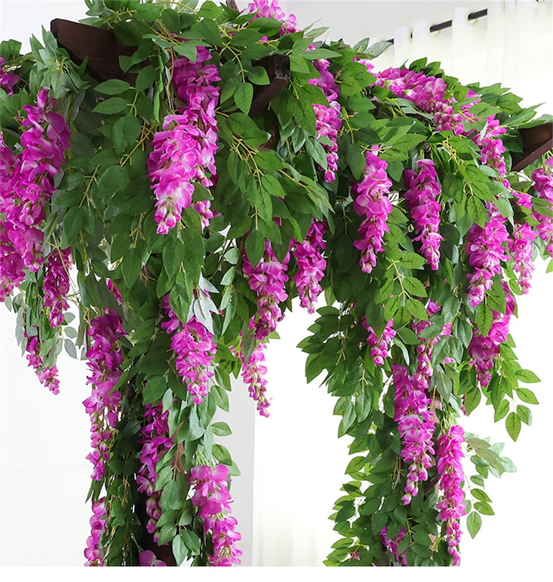 May include: Artificial wisteria garland with cascading purple flowers and green leaves. The garland is draped over a dark brown wooden structure, creating a vibrant and decorative display. Ideal for adding a touch of nature to any space.