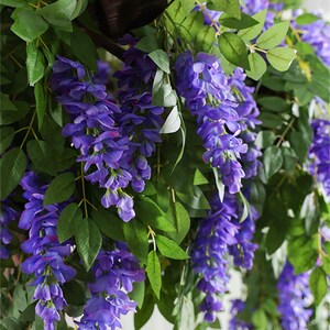 May include: Artificial wisteria vines with cascading purple flowers and green leaves. The faux floral arrangement is designed for decorative purposes, adding a touch of nature to interior spaces. The wisteria is a popular choice for weddings and home decor.