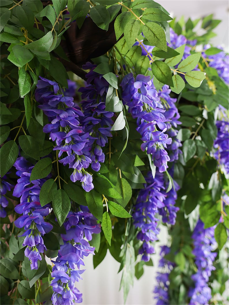 May include: Artificial wisteria vines with cascading purple flowers and green leaves. The faux floral arrangement is designed for decorative purposes, adding a touch of nature to interior spaces. The wisteria is a popular choice for weddings and home decor.