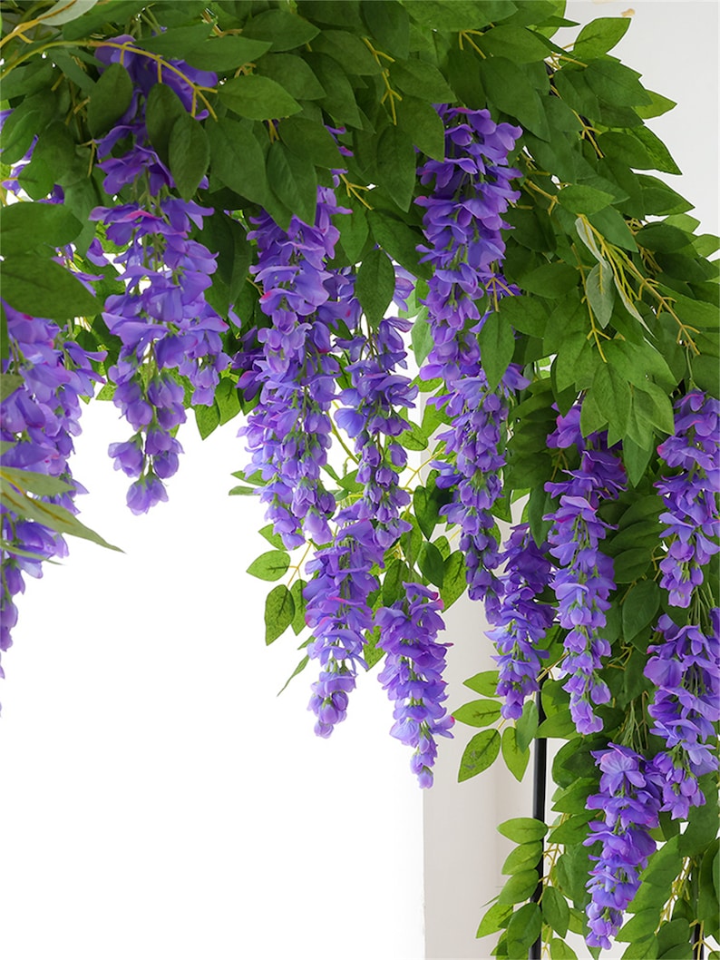 May include: Artificial wisteria vines with cascading purple flowers and green leaves. The wisteria is a popular decorative element for weddings, parties, and home decor. The image shows a close-up of the wisteria, highlighting the details of the flowers and leaves.