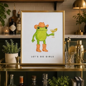 May include: A framed print on a gold bar cart featuring a green frog in a cowboy hat and boots holding a cocktail. The text "LET'S GO GIRLS" is below the image. The cart also holds liquor bottles, cocktail books, and bar tools.
