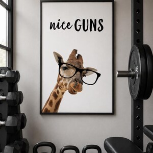 May include: A framed print featuring a giraffe wearing glasses with the text "nice GUNS" in black. The image is set in a gym with weights and a bench in the background. The giraffe has a brown and tan pattern.