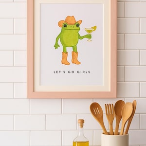 May include: A framed print featuring a green frog wearing a cowboy hat and boots, holding a cocktail. The text "LET'S GO GIRLS" is below the frog. The frame is light pink. Kitchen utensils and a bottle of oil are in the foreground.