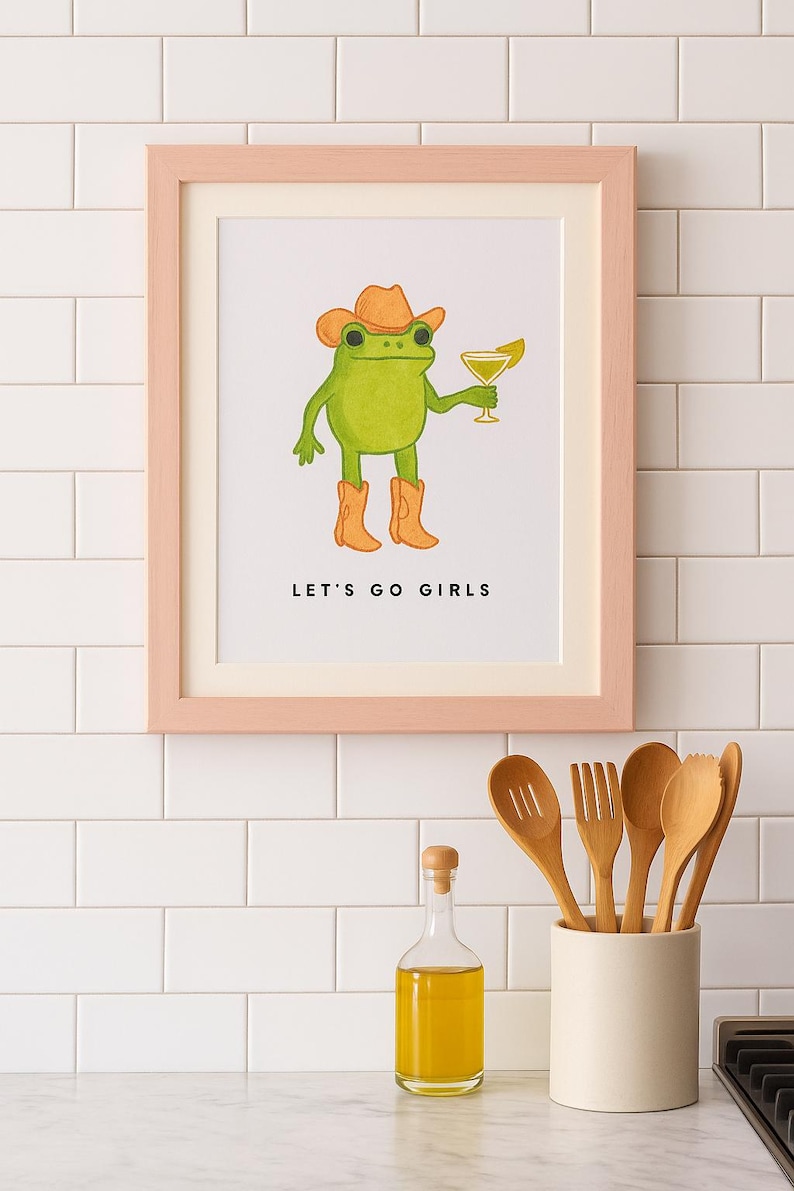 May include: A framed print featuring a green frog wearing a cowboy hat and boots, holding a cocktail. The text "LET'S GO GIRLS" is below the frog. The frame is light pink. Kitchen utensils and a bottle of oil are in the foreground.