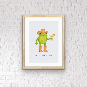 May include: A framed art print featuring a green frog wearing a cowboy hat and boots, holding a cocktail. The text "LET'S GO GIRLS" is printed below the frog. The artwork is set against a white background.