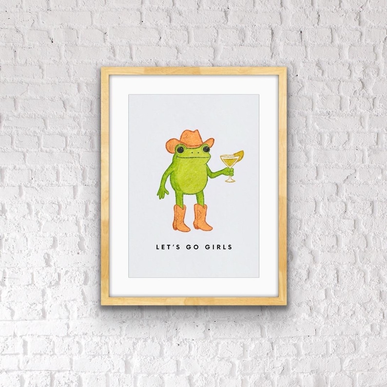 May include: A framed art print featuring a green frog wearing a cowboy hat and boots, holding a cocktail. The text "LET'S GO GIRLS" is printed below the frog. The artwork is set against a white background.