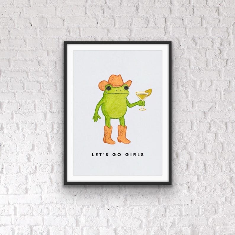 May include: A framed print featuring a green frog wearing a cowboy hat and boots, holding a cocktail. The phrase "LET'S GO GIRLS" is printed below the frog illustration. The artwork is set against a white background.