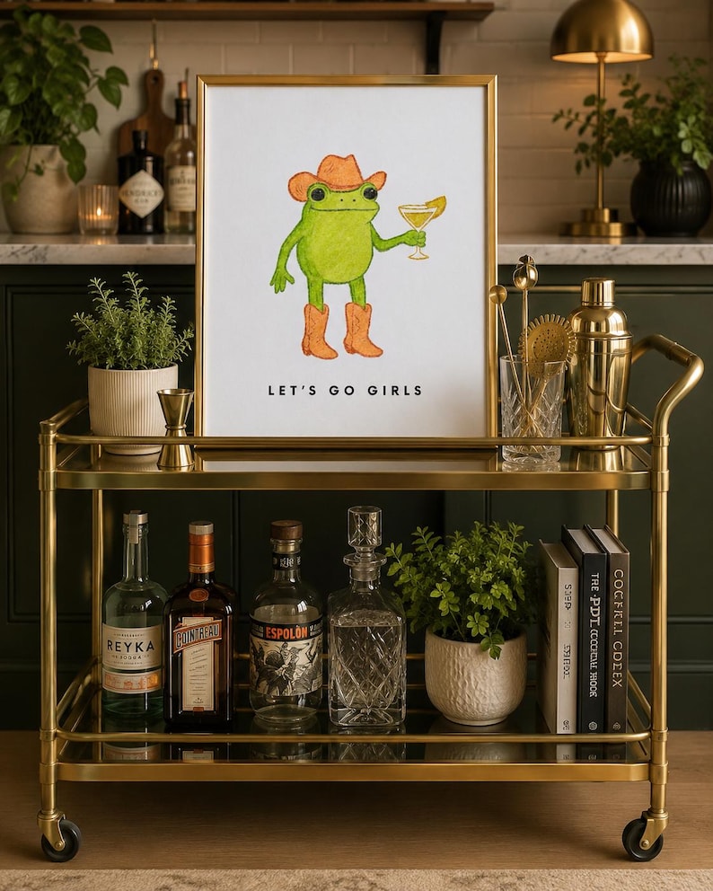 May include: A framed print on a gold bar cart featuring a green frog in a cowboy hat and boots holding a cocktail. The text "LET'S GO GIRLS" is below the image. The cart also holds liquor bottles, cocktail books, and bar tools.